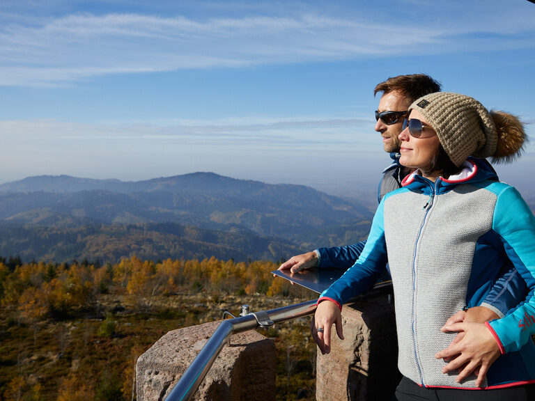 Ein Paar blickt nach dem geschafften Aufstieg erfüllt von einem Aussichtspunkt über den mittleren Schwarzwald.