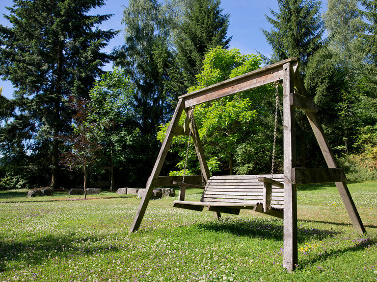 Ein rustikales Holzschaukel steht frei im Gras, eingerahmt von einem dichten Wald im weitläufigen Garten des Waldhotels Grüner Baum.