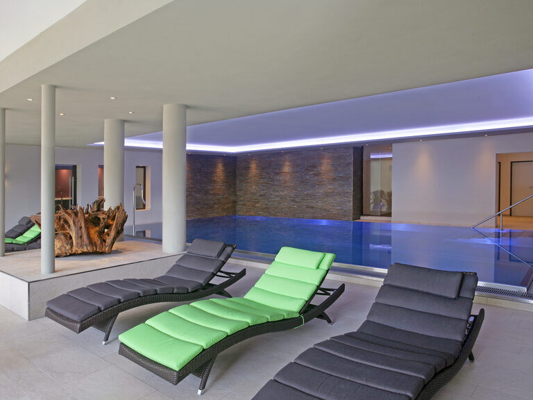 Eine moderne Poollandschaft im inneren des Wellnesshotels mit gemütlichen Liegen und einer stimmigen Beleuchtung.